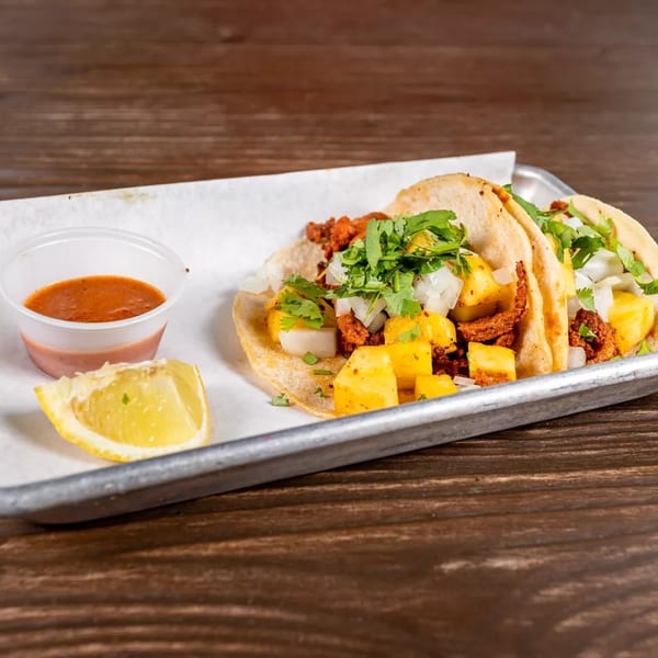 Two al pastor tacos with pineapple, onions, and cilantro, served with salsa and a lime wedge.