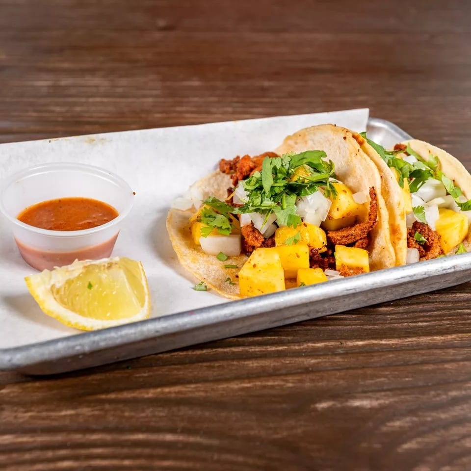 Two al pastor tacos with pineapple, onions, and cilantro, served with salsa and a lime wedge.
