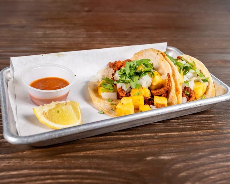 Two al pastor tacos with pineapple, onions, and cilantro, served with salsa and a lime wedge.