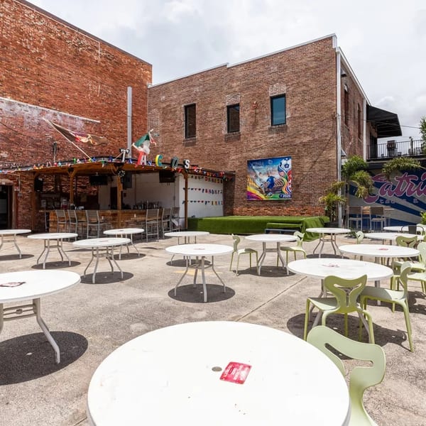 Outdoor patio with white tables and light green chairs, a bar, and brick buildings.