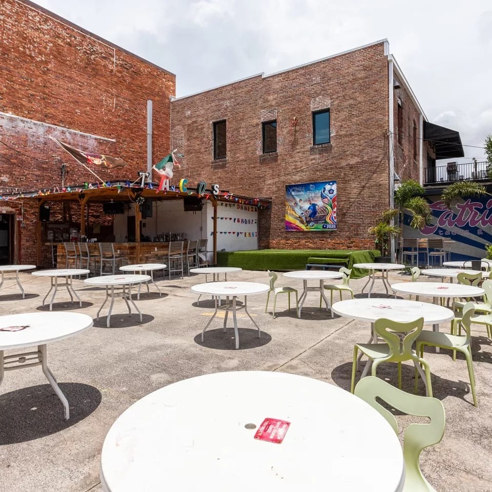 Outdoor patio with white tables and light green chairs, a bar, and brick buildings.