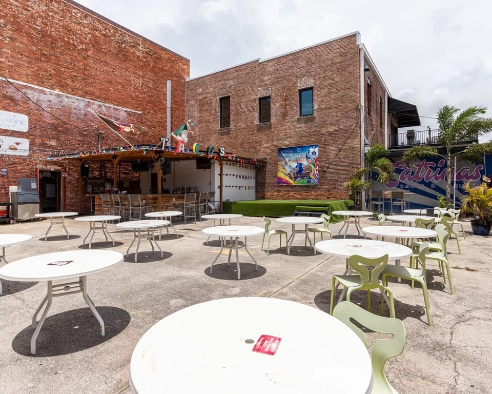 Outdoor patio with white tables and light green chairs, a bar, and brick buildings.
