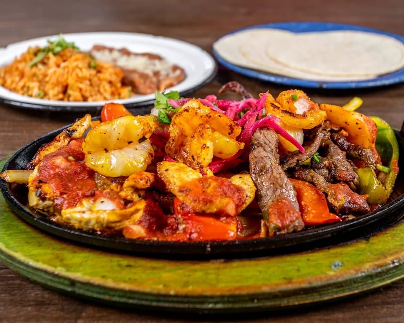 Sizzling fajitas with shrimp, steak, chicken, peppers, and onions, served with rice and tortillas.