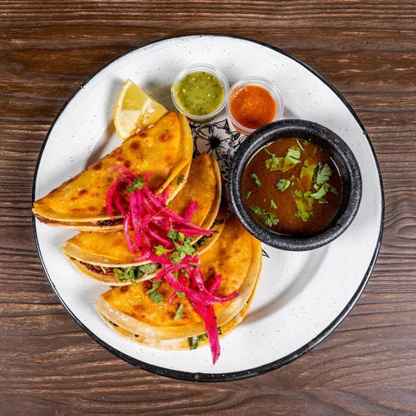 Three golden tacos topped with red pickled onions, served with dipping sauces and a lime wedge.