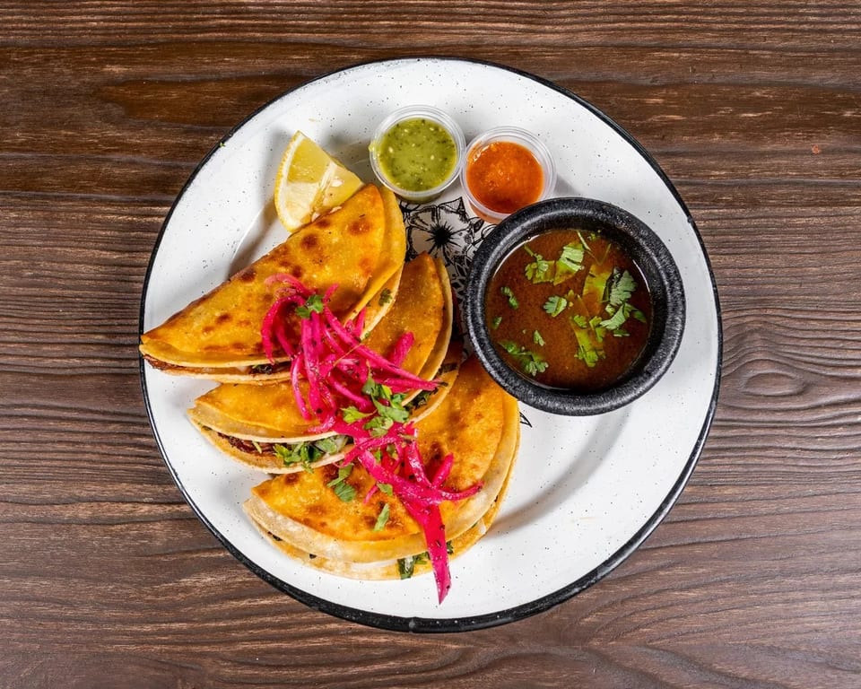 Three golden tacos topped with red pickled onions, served with dipping sauces and a lime wedge.