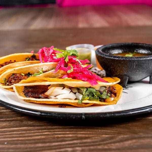 Three birria tacos with red pickled onions and cilantro, served with a side of consommé.