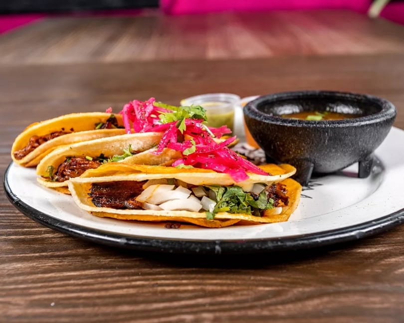 Three birria tacos with red pickled onions and cilantro, served with a side of consommé.