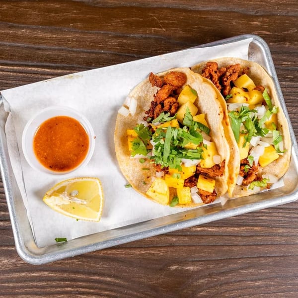 Two al pastor tacos with pineapple, onions, and cilantro, served with salsa and a lime wedge.