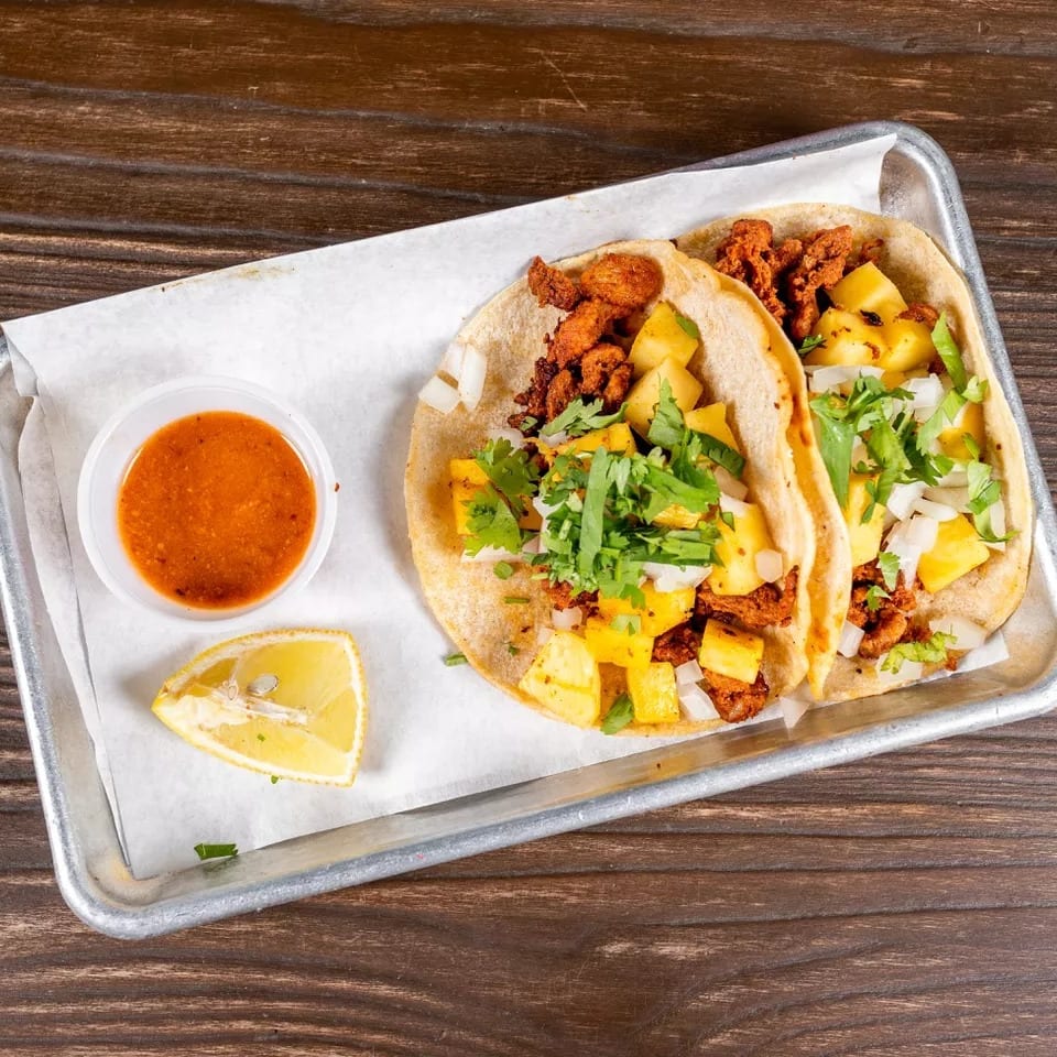 Two al pastor tacos with pineapple, onions, and cilantro, served with salsa and a lime wedge.