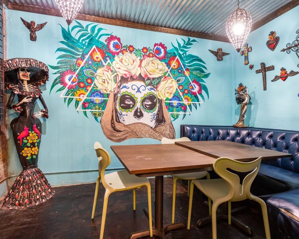 Restaurant interior with a vibrant Day of the Dead mural, blue booth, and light green chairs.