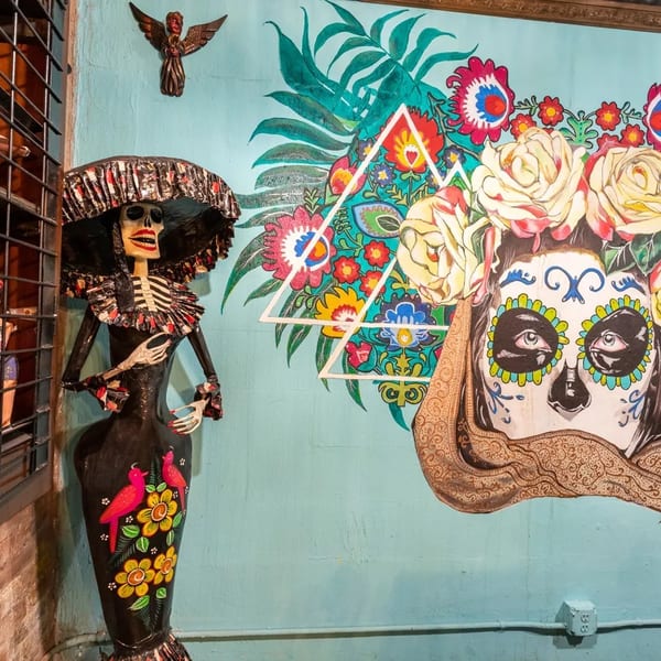 A Catrina skeleton doll stands next to a vibrant mural of a sugar skull with flowers.