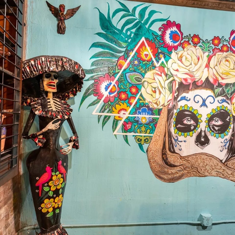 A Catrina skeleton doll stands next to a vibrant mural of a sugar skull with flowers.