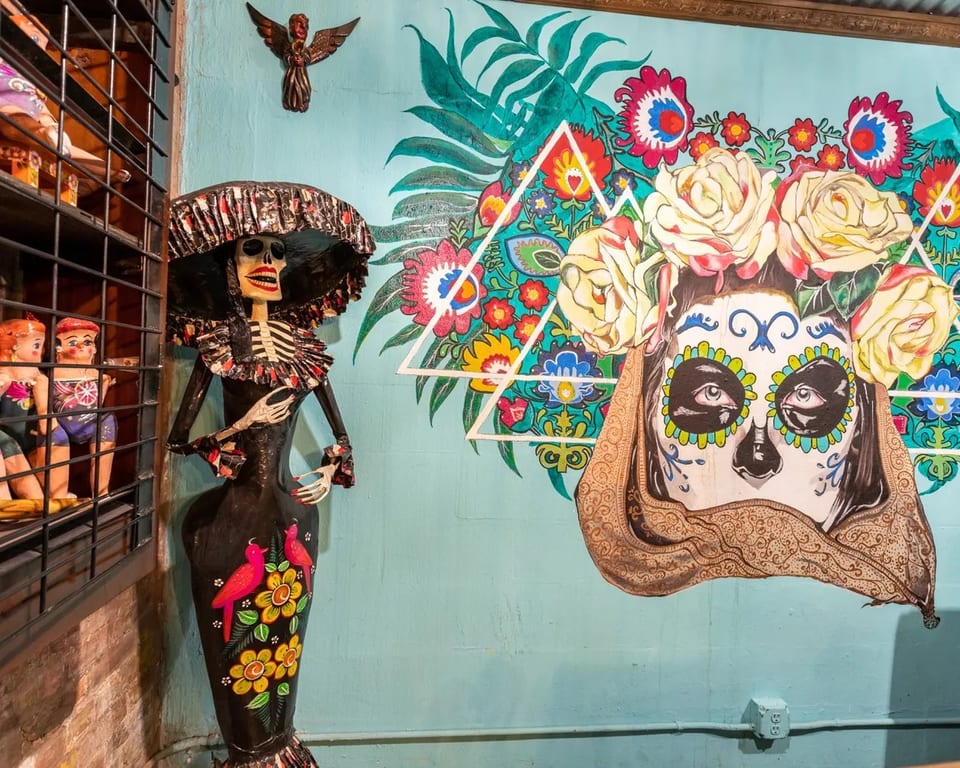 A Catrina skeleton doll stands next to a vibrant mural of a sugar skull with flowers.